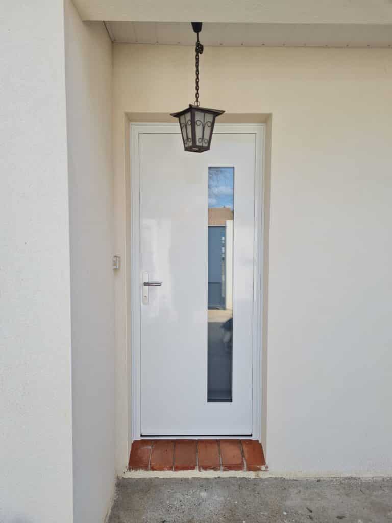 Porte d'entrée moderne blanche et laquée avec une longue fenêtre verticale, suspendue sous un luminaire extérieur en fer forgé.