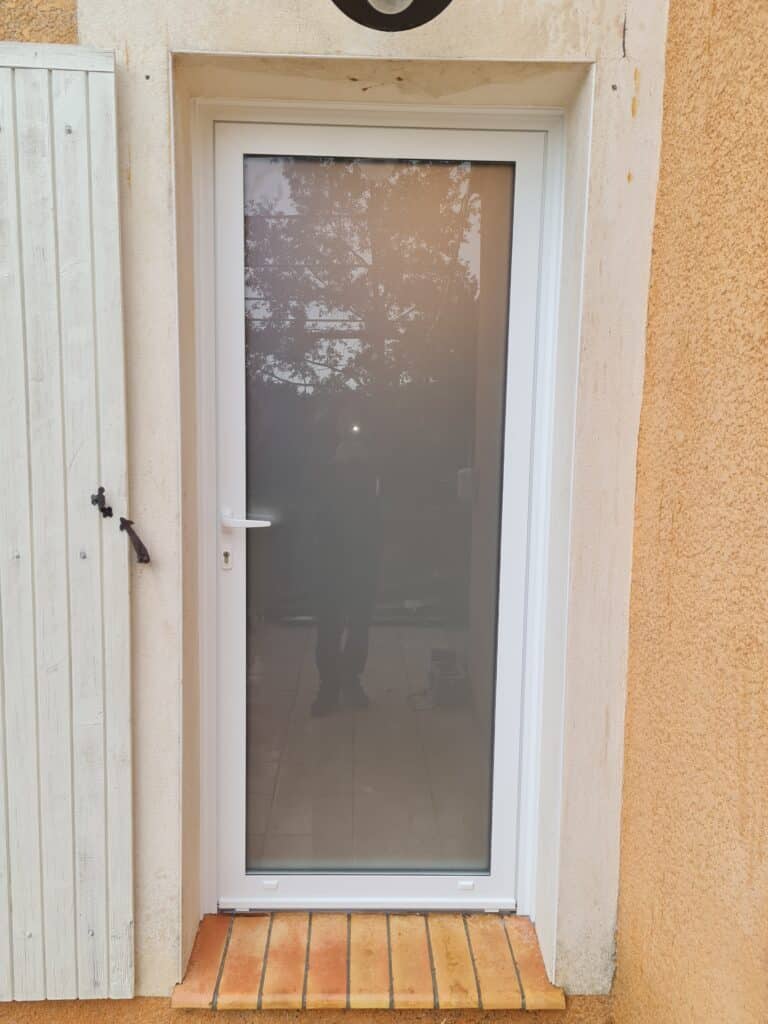 Porte vitrée PVC blanche neuve installée à côté d'un volet en bois blanc vieilli, sur mur ocre texturé, reflet d'une personne.