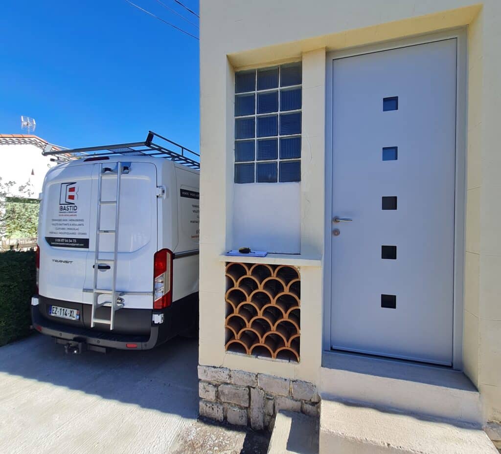 Pose menuiserie : Van BASTID et porte moderne Van BASTID menuiserie avec échelle, plaque EZ-114-XL, devant une maison avec porte moderne grise et ouverture d'aération en terre cuite.